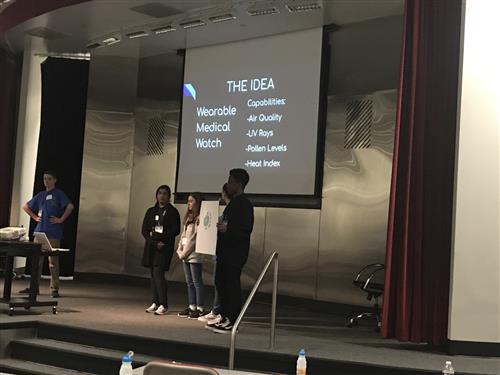 5 students stand on a stage presenting their idea with a screen of their presentation behind them. 